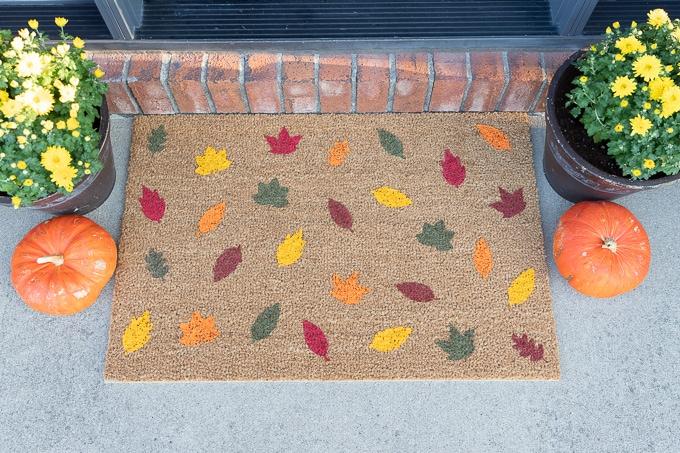 Image of doormat with leaves painted on it.  There are mums and pumpkins on each side of the mat.